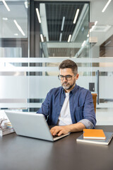 Obraz premium Middle-aged man with glasses and beard in a denim shirt concentrates on his silver laptop at a modern office desk, typing and planning digital work for his business