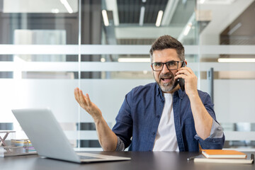 Obraz premium Mature businessman experiencing stress and frustration, shouting into his mobile phone while working at his office desk, indicating conflict and professional burnout