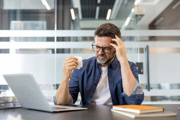 Obraz premium Stressed businessman touching his temples with one hand and holding a crumpled tissue in the other, experiencing a headache and symptoms of burnout while working at his desk in a modern office