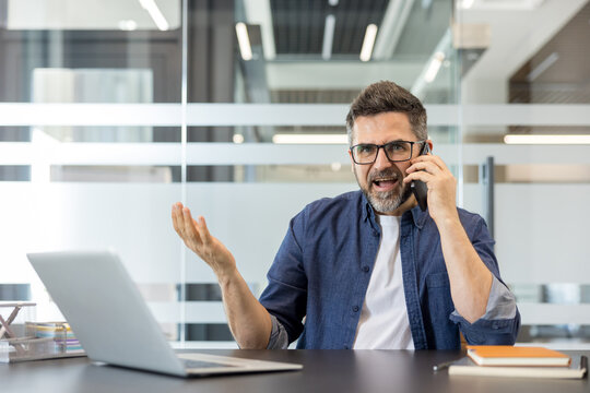 Mature businessman experiencing stress and frustration, shouting into his mobile phone while working at his office desk, indicating conflict and professional burnout