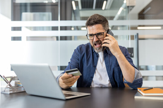 Stressed man having an online financial problem, sitting at office desk, holding a credit card and shouting while talking on the phone with customer support