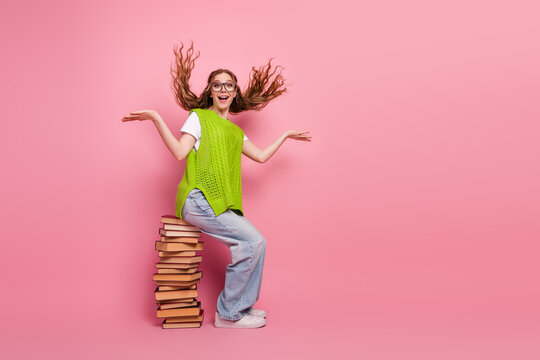 Happy young woman student wearing green vest sits on stack of book against pink background posing playfully at school style shot