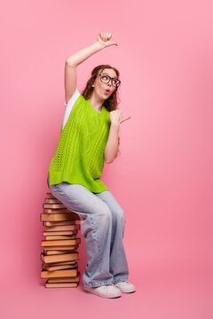 Young female student sits on a stack of book on a pink studio backdrop wearing a bright lime vest and glasses for a playful fashion stock image