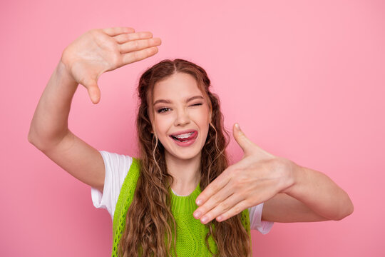 Playful teen girl framing a photo with her hands against a pink background wearing a green vest