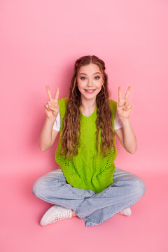 Young girl sits cross legged in bright green vest smiling and making peace signs against pink background