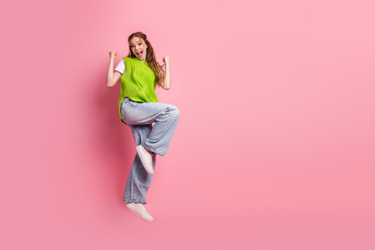 Young woman jumping joyful fashion moment wearing green vest jeans pink background smiling lively trendy style school day mood fun