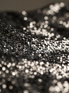 Close-up of a black fabric with small silver sequins scattered across it. the sequins are of different sizes and shapes, creating a textured and shiny surface.