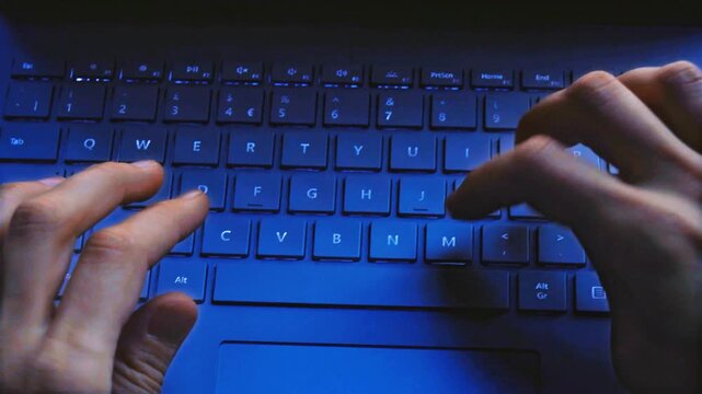 Close-up shot of hands typing fast on a blue backlit laptop keyboard in a dark room. Ideal for coding, programming, software development, data entry, cyber security, and technology-related content.