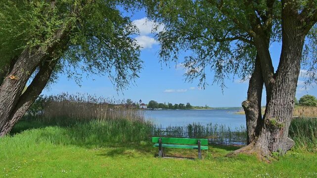 Eine gr&uuml;ne Parkbank am Ufer eines Sees mit Blick auf ein gegen&uuml;berliegendes Dorf, Rhodes, Etang du Stock, Sarrebourg, Moselle, Frankreich