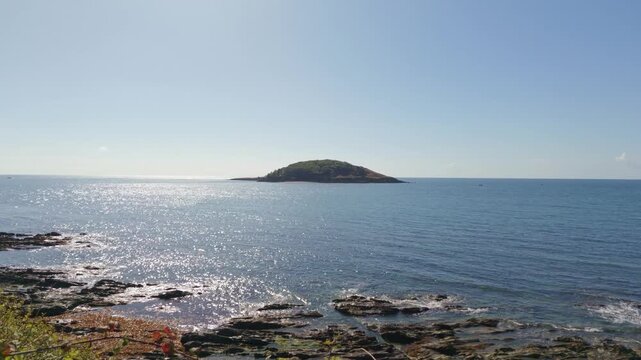 Static view from Hannafore shows Looe Island in the distance across a bright blue sea. Rocky terrain and vegetation line the foreground with waves gently lapping at the high tide mark along the shore
