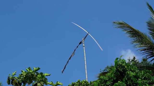 Traditional bamboo wind propeller spinning against clear blue sky in tropical garden, used as folk bird scarecrow or decorative garden art in Southeast Asian rural landscape.