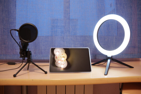 Podcast microphone and ring light beside tablet showing festive snowman image on wooden desk with blue curtain backdrop, creating cozy studio workspace still life