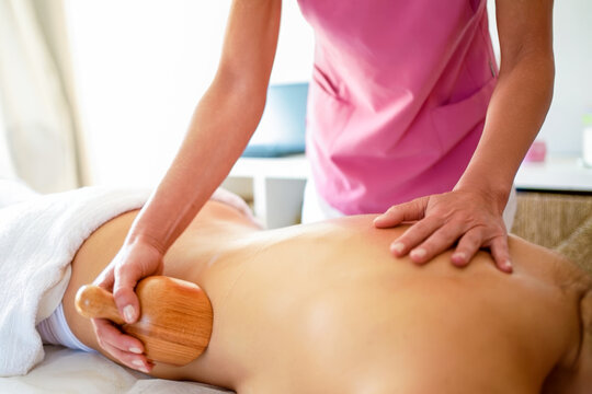 Massage therapist applying wooden cup treatment and hand pressure to bare back in bright wellness room with soft natural light, calm linens, professional bodywork