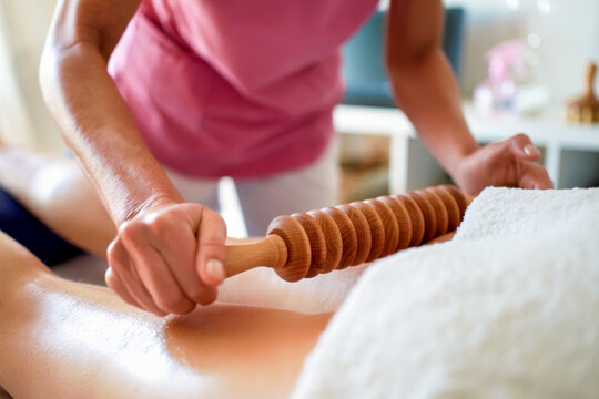 Therapist performing manual massage with wooden roller on bare back in bright treatment room, close view of spa bodywork with client resting face down on padded table