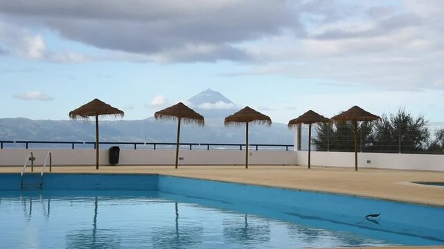 Thatched parasols beside an empty swimming pool with Mount Pico beyond, Sao Jorge Island, Azores, Portugal