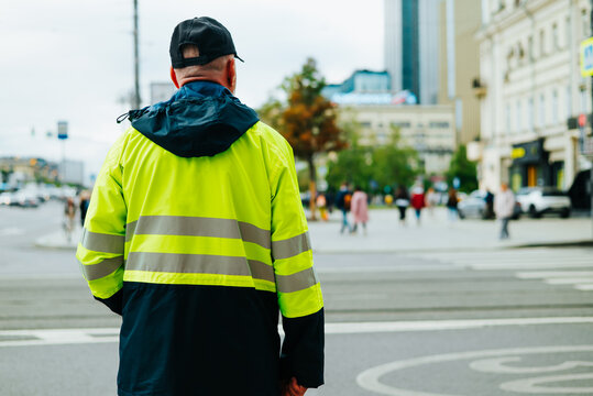 A traffic worker in a high-visibility jacket stands at a crosswalk observing the road. Pedestrians pass by in an urban setting with buildings visible in the background