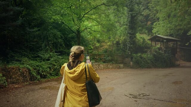 Rear View Of Woman In Yellow Jacket Photographing Forest Path With Phone
