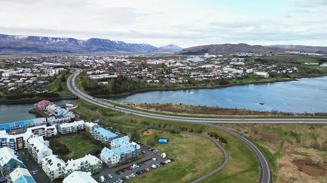 Aerial view of Reykjavik suburbs beside a lake, with curving roads, colorful buildings and Mount Esja rising across the horizon under soft cloudy light 4K.