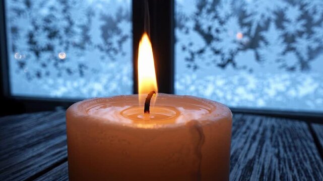 Burning candle with a warm flame in the foreground set against a frosty window with ice patterns evoking a cozy winter atmosphere