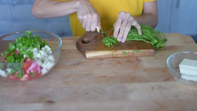 Woman chopping fresh dill on wooden cutting board while preparing healthy vegetable salad with feta cheese in kitchen