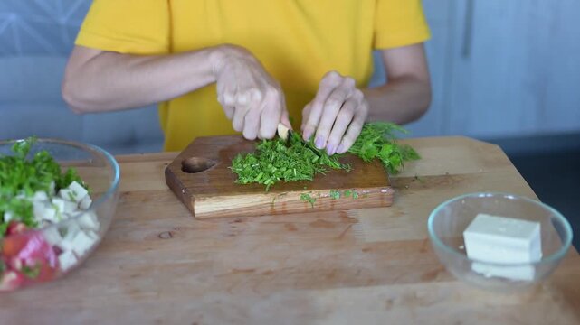 Woman chopping fresh dill on wooden cutting board while preparing healthy vegetable salad with feta cheese in kitchen