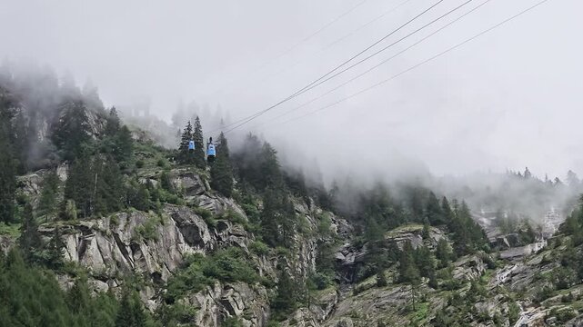 Blue cable cars above Staffa on the Monte Moro line, Macugnaga, Piedmont, Italy, climbing into thick mist over forested crags