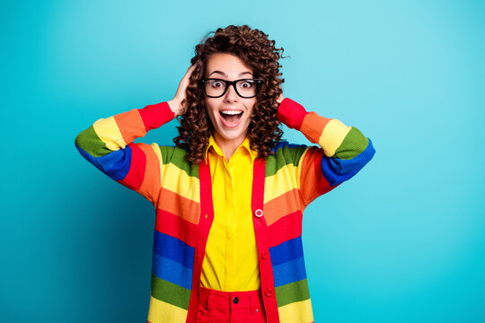 Happy young woman in glasses wearing a colorful rainbow sweater and bright yellow shirt, surprised and excited portrait against a turquoise background