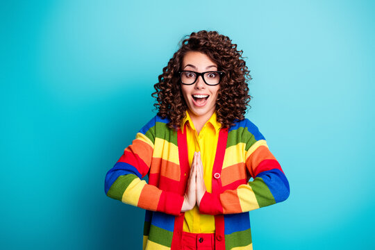 Excited young woman in rainbow sweater and glasses smiling with grateful hands against a turquoise background
