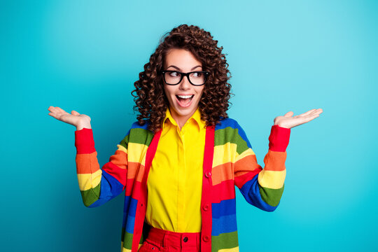 Cheerful young woman in rainbow cardigan and glasses gesturing with open palms against a bright turquoise background
