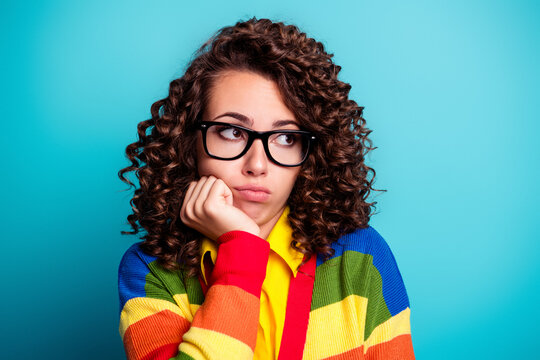 Thoughtful woman in glasses wearing a colorful striped sweater against a bright blue background, looking pensive and bored with hand on chin in a casual portrait