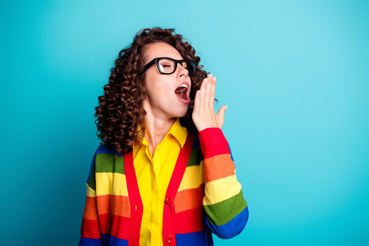Young woman yawning in colorful rainbow sweater and glasses against turquoise background, expressing tiredness, boredom and morning fatigue