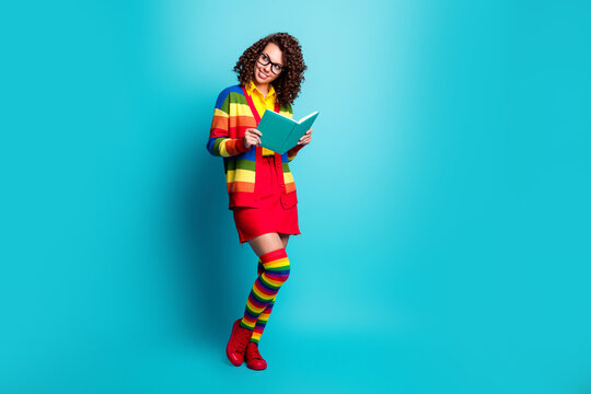 Young woman reading a book in colorful rainbow outfit and knee socks, smiling with glasses against a bright turquoise background