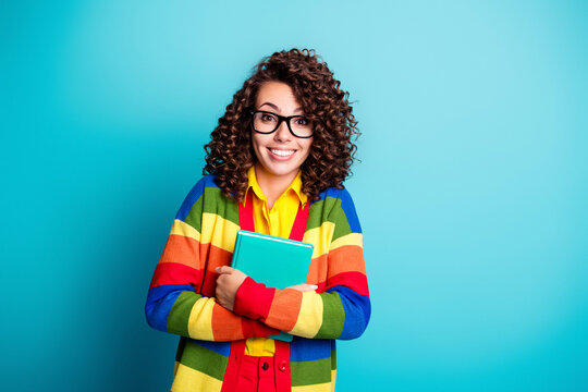 Smiling student woman in rainbow sweater holding notebook against turquoise background, cheerful young learner with glasses and curly hair