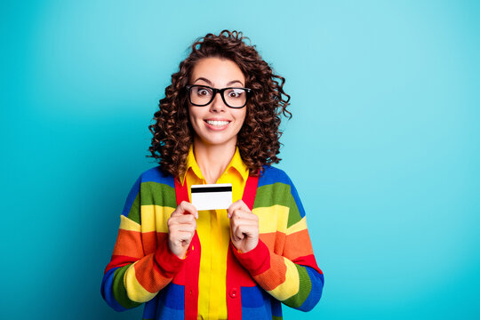 Happy young woman holding credit card in colorful rainbow sweater smiling against blue background