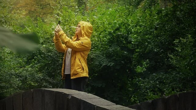 Woman In Yellow Raincoat Taking Photos With Smartphone In Forest