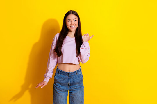 Young woman smiling and pointing to the side on a bright yellow background wearing a cropped pink top and denim jeans