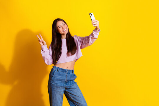 Young woman taking a selfie with smartphone and peace sign on bright yellow background, smiling and casual fashion portrait