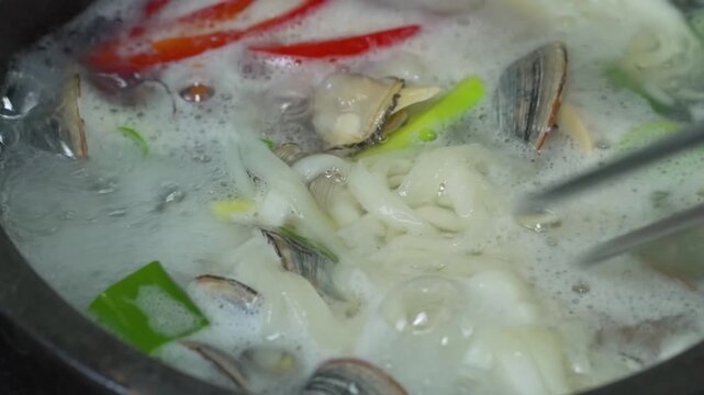 Seafood stew simmering in pot with red chilies onion greens foamy broth, warm home kitchen mood, with seafood, stew, clam, mussel detail, showing shell, shellfish, red, chili context