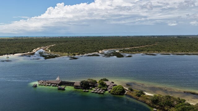 Aerial view of Parque Nacional dos Lencois Maranhenses featuring a small island with thatched-roof structures surrounded by clear water and lush vegetation in Atins, Maranhao, Brazil.