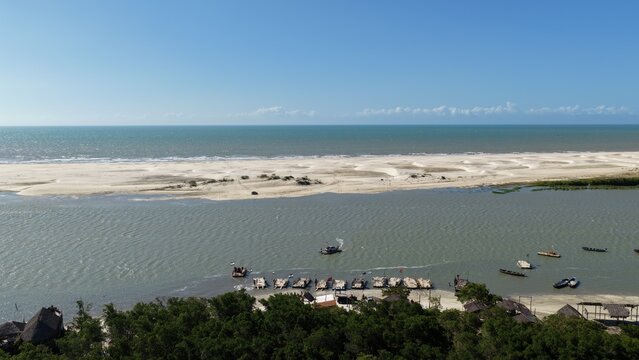Aerial view of Parque Nacional dos Lencois Maranhenses showing the river meeting the sea, with wooden boats moored along the sandy bank, dunes, and vegetation in Atins, Maranhao, Brazil.