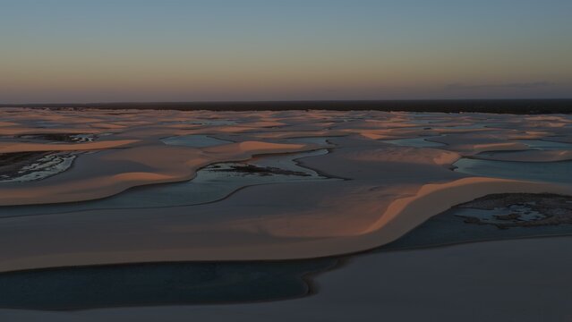 Aerial view of Parque Nacional dos Lencois Maranhenses with vast white sand dunes and seasonal rainwater lagoons under a soft sunset sky in Atins, Maranhao, Brazil.