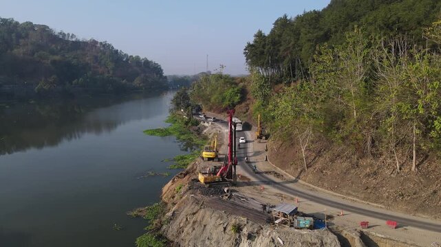 Aerial view of road construction site with heavy machinery and vehicles along the riverbank in Banyumas, Central Java, Indonesia.
