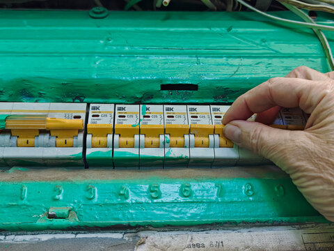 Kirov, Russia - August 20, 2026: Hand grips a yellow IEK C25 circuit breaker lever in an old green-painted electrical cabinet with a row of numbered automatic switches.