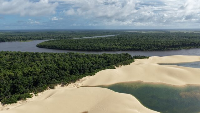 Aerial view of Parque Nacional dos Lencois Maranhenses featuring vast white sand dunes and turquoise lagoons bordering a lush green forest in Atins, Maranhao, Brazil.