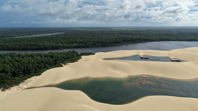 Aerial view of Parque Nacional dos Lencois Maranhenses featuring vast white sand dunes, crystal clear lagoons, and a lush river landscape in Atins, Maranhao, Brazil.