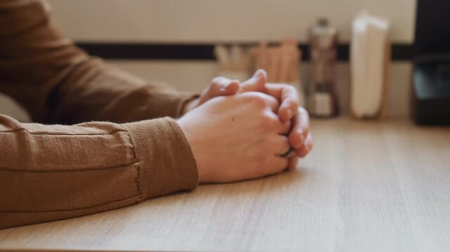 Close up of person clasped hands resting on light wood surface with soft natural lighting. Background includes blurred objects like brushes and containers suggesting indoor workspace