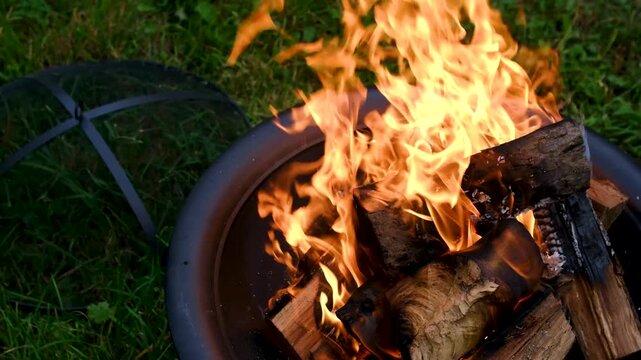 Dry logs burn with strong orange flames inside a round metal fire pit outdoors, creating heat and glowing charcoal for grilling and summer cooking. Close up.