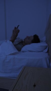 Man trying to sleep but reaching for smartphone to scroll again, compulsive phone use and insomnia at night with selective focus, vertical