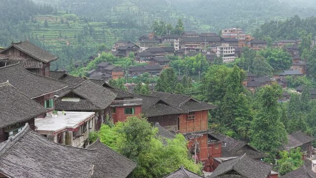 Aerial view of traditional wooden houses nestled among lush green terraced hills and dense forest in Qiaoniao Village, Guizhou, China.