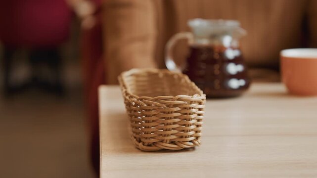 Close up of person hands placing and adjusting small wicker basket on wooden table. scene includes soft lighting with visible objects like glass coffee carafe in background. focus remains